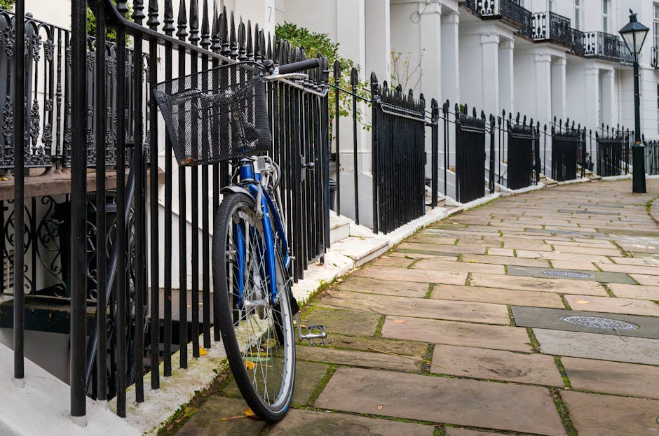 A black metal fence lining a residential sidewalk with irregularly shaped stone paving, next to a white building with columns. A blue bicycle with a basket is locked to the fence, which appears clean and well-maintained. The pavement is clear and free of debris, with some moss or grass growing between the stones. The scene is outdoors with natural lighting, and the overall environment looks tidy and cared for, consistent with deep surface cleaning or maintenance as promoted by carpetcleanerssouthkensington.co.uk for avoiding hidden cleaning charges in South Kensington.