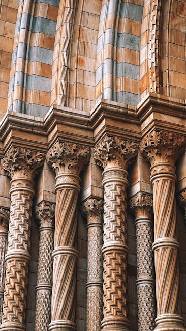 Close-up view of a detailed section of Gothic-style stone columns with ornate capitals and intricate carvings. The columns feature a mix of light brown and beige hues, with some darker accents highlighting the decorative patterns. The stone surfaces exhibit a clean, polished appearance with no visible dust or dirt. Above the columns, part of the stone wall shows layered structural elements with a combination of smooth and textured finishes, illuminated by soft, natural light. This architectural detail is likely located in a historic church or cathedral setting, emphasizing craftsmanship and meticulous surface cleaning by professionals like those at carpetcleanerssouthkensington.co.uk to maintain the integrity and appearance of such surfaces.