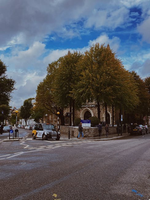 A street scene outside a historic building with a prominent arched entrance, surrounded by large, leafy trees with green and brown foliage, indicating autumn. The pavement and road surface are clean and dry, with clear pedestrian crossings and traffic signals visible. Several parked and moving cars, a few pedestrians, and street lamps are present in the scene. Natural daylight illuminates the setting, highlighting the weathered brickwork of the building and the vibrant sky overhead with scattered clouds. This image captures an urban environment in South Kensington, suitable for illustrating the importance of routine cleaning and maintenance in a city setting, as promoted by carpetcleanerssouthkensington.co.uk for end of tenancy cleaning and surface sanitisation.