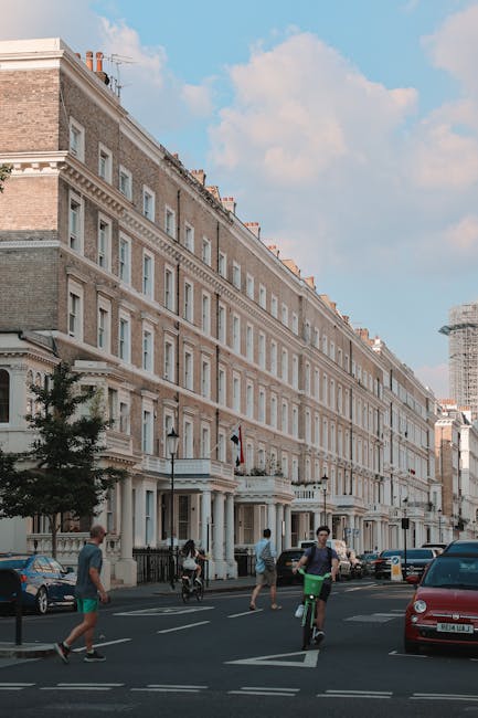 Exterior view of a row of charming commercial buildings on Exhibition Road South Kensington, showcasing large glass display windows and decorative balconies above. The building facades feature pastel green and white paint with detailed trim and wrought iron balcony railings. The sidewalk in front is clean, with a small blackboard sign and potted plants near the entrance of a flower shop. A street sign and bicycle lane markings are visible on the asphalt road, which appears well-maintained and free of debris, highlighting a tidy urban environment in South Kensington. This setting may be the exterior of a retail or gallery space, emphasizing the importance of surface cleaning and maintenance for pristine curb appeal, as provided by [COMPANY_NAME].