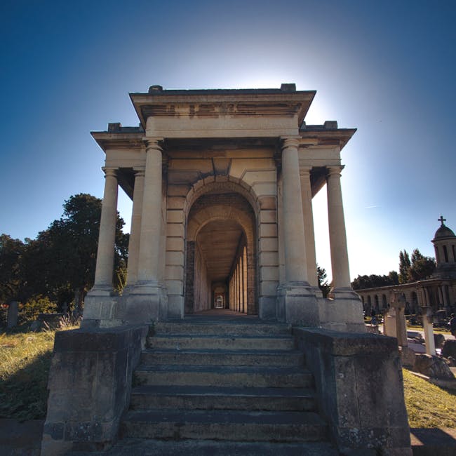 A stone mausoleum with classical architectural features including large, round-columned portico and stone steps leading up to an arched entrance, situated in a sunlit cemetery with nearby trees and other memorial structures visible in the background, under a clear blue sky.