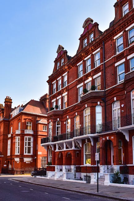 A row of Victorian-style red brick residential buildings on Old Brompton Road in South Kensington, featuring large sash windows with white trims and decorative wrought iron balconies. The buildings have multiple stories, with some windows open and others closed, and the street below is clean and well-maintained. Bright daylight illuminates the facades against a clear blue sky, highlighting the architectural details and the pristine appearance of the exteriors, indicative of regular surface cleaning and maintenance by professional cleaning services such as those offered by carpetcleanerssouthkensington.co.uk.