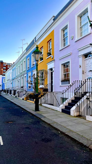 Image of a row of colorful terraced houses along a residential street in South Kensington, with bright blue, yellow, and purple facades. The houses feature white-framed windows, black wrought-iron railings, and small front gardens accessible via flight stairs. A vintage-style street lamp stands near the sidewalk, illuminated by daylight. The scene depicts a clean, well-maintained urban environment with tidy pavement and no visible debris, illustrating the importance of surface cleaning and maintenance for overall hygiene in residential areas. [COMPANY_NAME], a professional cleaning service, emphasizes the significance of deep cleaning and sanitisation for property upkeep, especially relevant in the context of same-day cleaning delays in South Kensington.