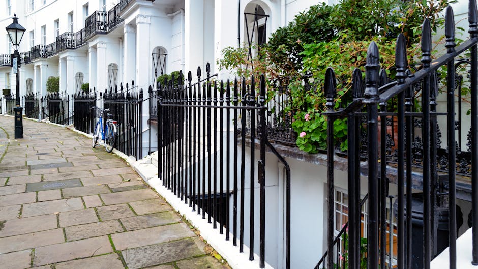 A paved sidewalk in South Kensington lined with black wrought iron railings enclosing residential building entrances. The sidewalk consists of rectangular stone slabs, some of which are slightly uneven and stained, with patches of moss in the gaps. Behind the railings, white-painted building facades feature large black-framed windows and small balconies, with greenery and flowering plants adding touches of natural color. A blue bicycle is parked against the railings. The scene is illuminated by natural daylight, highlighting the clean, well-maintained appearance of the area, which is indicative of professional cleaning and surface maintenance as emphasized by [COMPANY_NAME] in their domestic and commercial cleaning services.