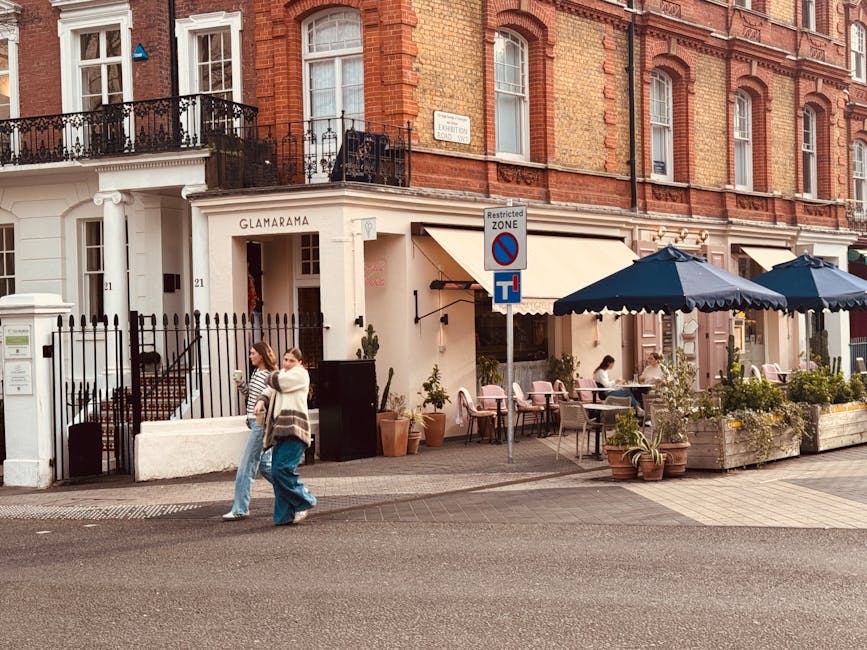 Exterior view of a restaurant with a white facade and a sign reading 'Glamarama' on Gloucester Road in South Kensington. The sidewalk features outdoor seating with several tables shaded by large dark blue umbrellas, and potted plants and flowers are arranged around the seating area. Two women are walking along the pavement, engaged in conversation. The scene is illuminated by natural daylight, highlighting the clean, well-maintained appearance of the outdoor space. The building has decorative black iron railings and large windows, contributing to an elegant urban atmosphere. This image emphasizes the importance of maintaining outdoor surfaces and seating areas through professional cleaning and sanitisation, as provided by companies like Carpet Cleaners South Kensington.